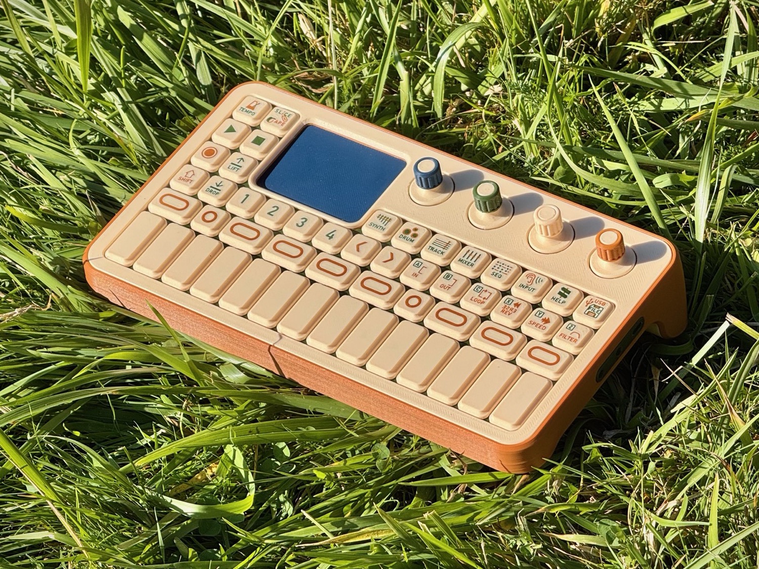 The Arbutus look-alike model lying in grass: a beige keyboard with an orange-brown base and four coloured knobs