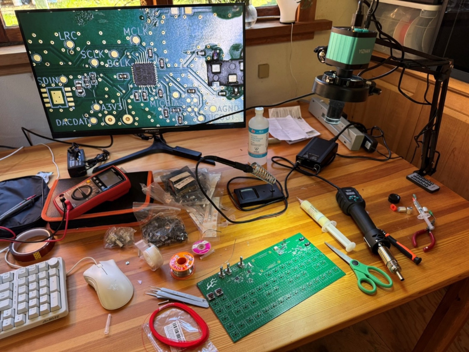 Desk with a microscope camera on an arm, a monitor showing a magnified circuit board, a multimeter, hot air station and tools
