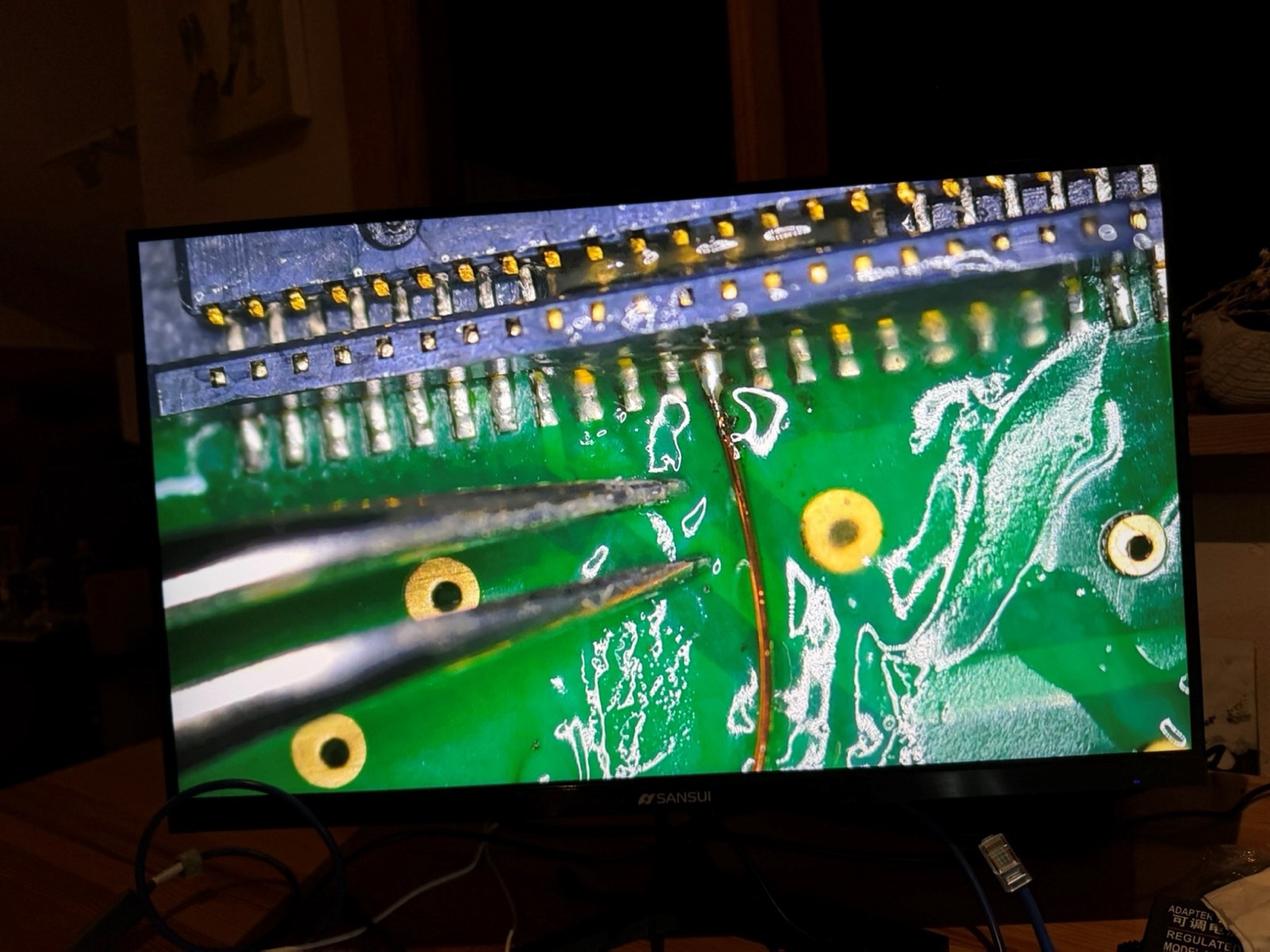 Monitor showing a magnified view of tweezers placing a hair-thin copper wire onto a pin of a 100-pin connector